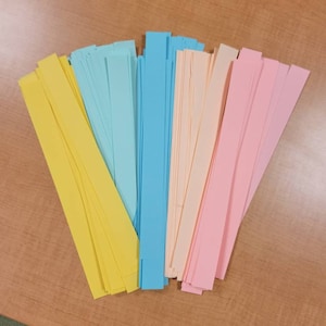 May include: A collection of colorful paper strips in yellow, light blue, peach, and pink. The strips are arranged in a fan shape on a wooden surface.