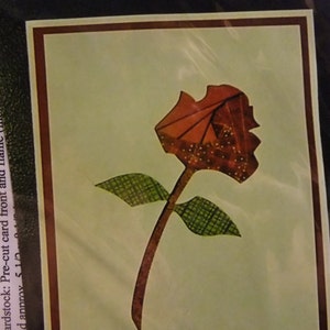 May include: A red paper rose with green leaves on a light green background. The rose is made of paper and has a textured look. The leaves are also made of paper and have a checkered pattern.