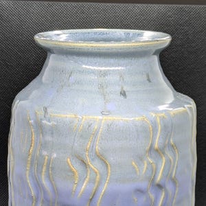 May include: A handcrafted ceramic vase with a blue and lavender glaze. The vase features a textured surface with vertical, wavy, golden-colored lines. The vase has a rounded body and a slightly flared rim, suitable for home decor.