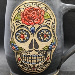 May include: A black ceramic mug with a sugar skull design. The skull is decorated with a red rose, blue and yellow flowers, and gold accents.