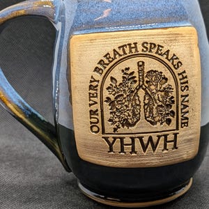 May include: Blue and brown ceramic mug with a handle. The mug features a rectangular, light brown patch with the engraved text "OUR VERY BREATH SPEAKS HIS NAME" and "YWHW". The patch also has a floral and lung design.
