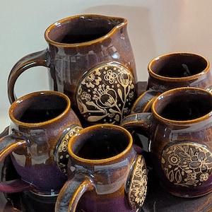 May include: A brown ceramic tea set with a pitcher, four mugs, and a tray. The set features a floral design in gold. The mugs and pitcher have a dark purple interior. The set is designed for serving tea or coffee.