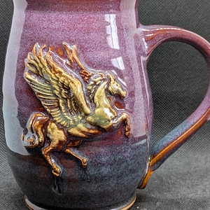 May include: A ceramic mug with a glossy finish that transitions from a deep purple at the top to a dark blue at the base. A raised, three-dimensional relief of a winged horse in a metallic gold and bronze tone is centered on the front surface.