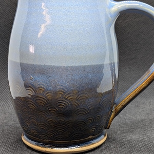 May include: A ceramic mug with a blue and brown glaze. The upper portion is light blue, transitioning to a darker blue and brown at the base. The bottom section features a textured, wave-like pattern. The handle is also blue and brown.