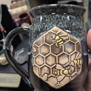 May include: A ceramic mug with a dark brown base and a mottled blue-grey rim. A raised hexagonal honeycomb relief is centered on the front, featuring two sculpted bees with yellow-painted bodies and brown wings against the textured cells.