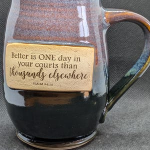 May include: A handcrafted ceramic mug with a brown and black glaze. The mug features a rectangular plaque with the quote "Better is ONE day in your courts than thousands elsewhere" and the reference "PSALM 84:10".