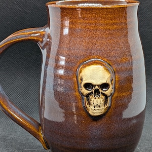 May include: A brown ceramic mug with a skull design. The mug has a large handle and a glossy finish. The skull is gold-colored and is centered on the mug. The mug is approximately 5 inches tall.