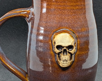 May include: A brown ceramic mug with a skull design. The mug has a large handle and a glossy finish. The skull is gold-colored and is centered on the mug. The mug is approximately 5 inches tall.