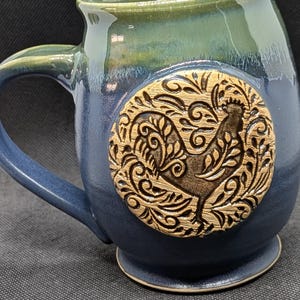 May include: A ceramic mug featuring a two-tone glaze with a dark blue base and a green rim. A circular medallion is centered on the front, displaying a raised, textured relief of a rooster surrounded by intricate, swirling floral patterns.