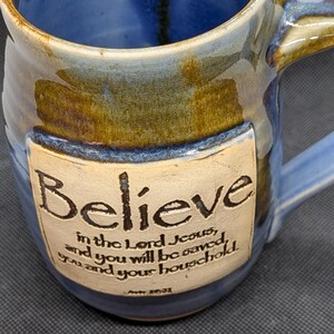 May include: Blue ceramic mug with a handle, featuring the word "Believe" and a religious quote. The mug has a blue interior and a brown and blue exterior, with a cream-colored label. A decorative piece for the home.