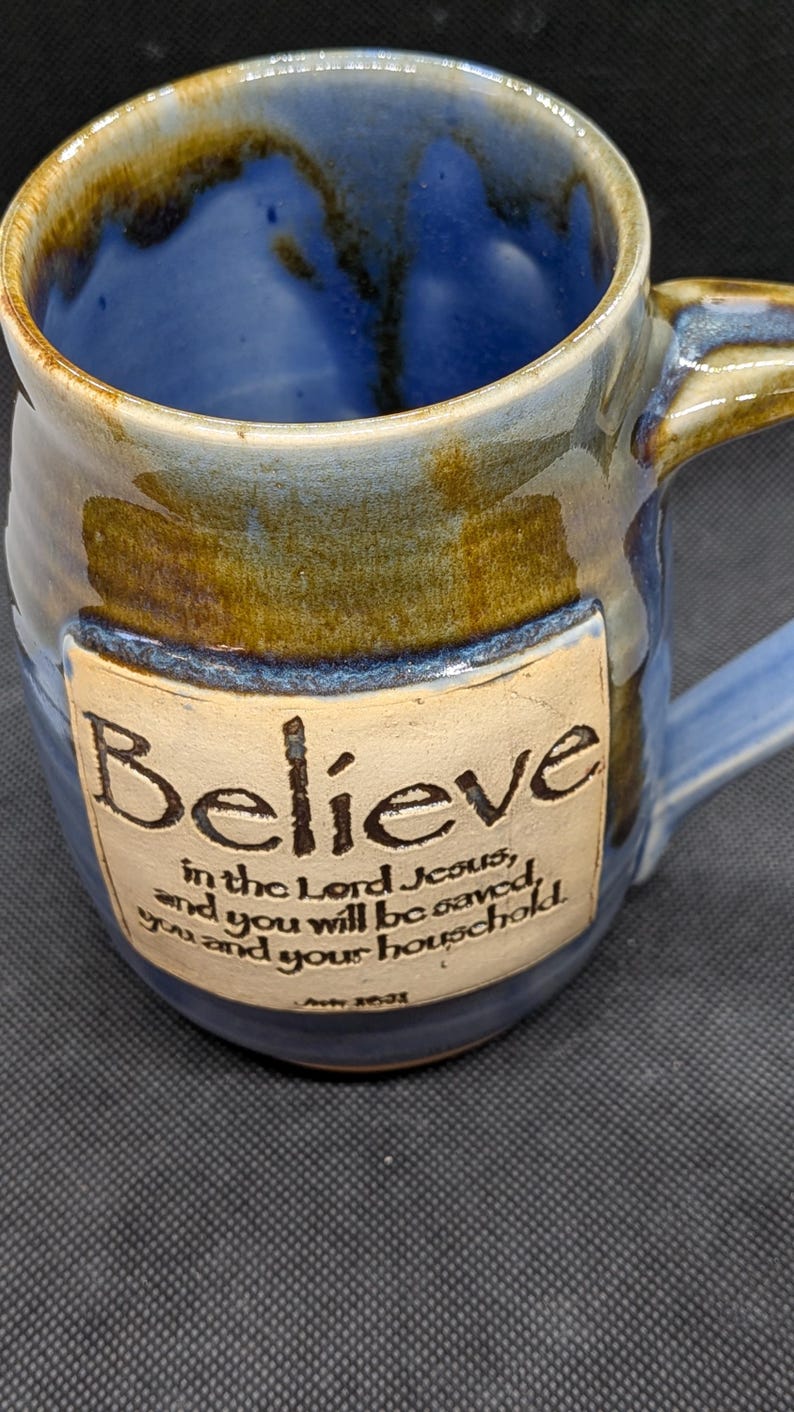May include: Blue ceramic mug with a handle, featuring the word "Believe" and a religious quote. The mug has a blue interior and a brown and blue exterior, with a cream-colored label. A decorative piece for the home.