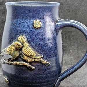 May include: A ceramic mug with a deep blue glaze. The mug features a raised relief design of two birds on a branch in shades of gold and brown. A small floral design is also present. The mug has a curved handle.