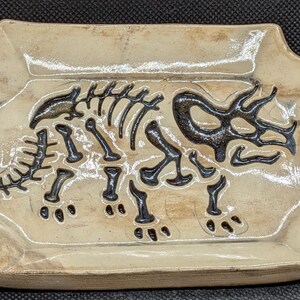 May include: A rectangular ceramic tray with beveled edges, featuring a glossy cream-colored glaze with brown mottled accents. The center of the tray displays a dark brown, recessed graphic of a triceratops dinosaur skeleton in profile.