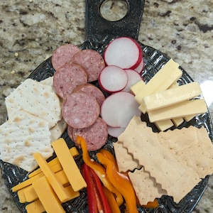 May include: A black ceramic serving board with a variety of snacks, including sliced sausage, crackers, cheese sticks, radishes, and red and yellow bell peppers.
