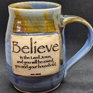 May include: A handcrafted ceramic mug with a blue and brown glaze. The mug features the word "Believe" and a Bible verse. The handle is a curved, sturdy design. A unique piece for coffee or tea.