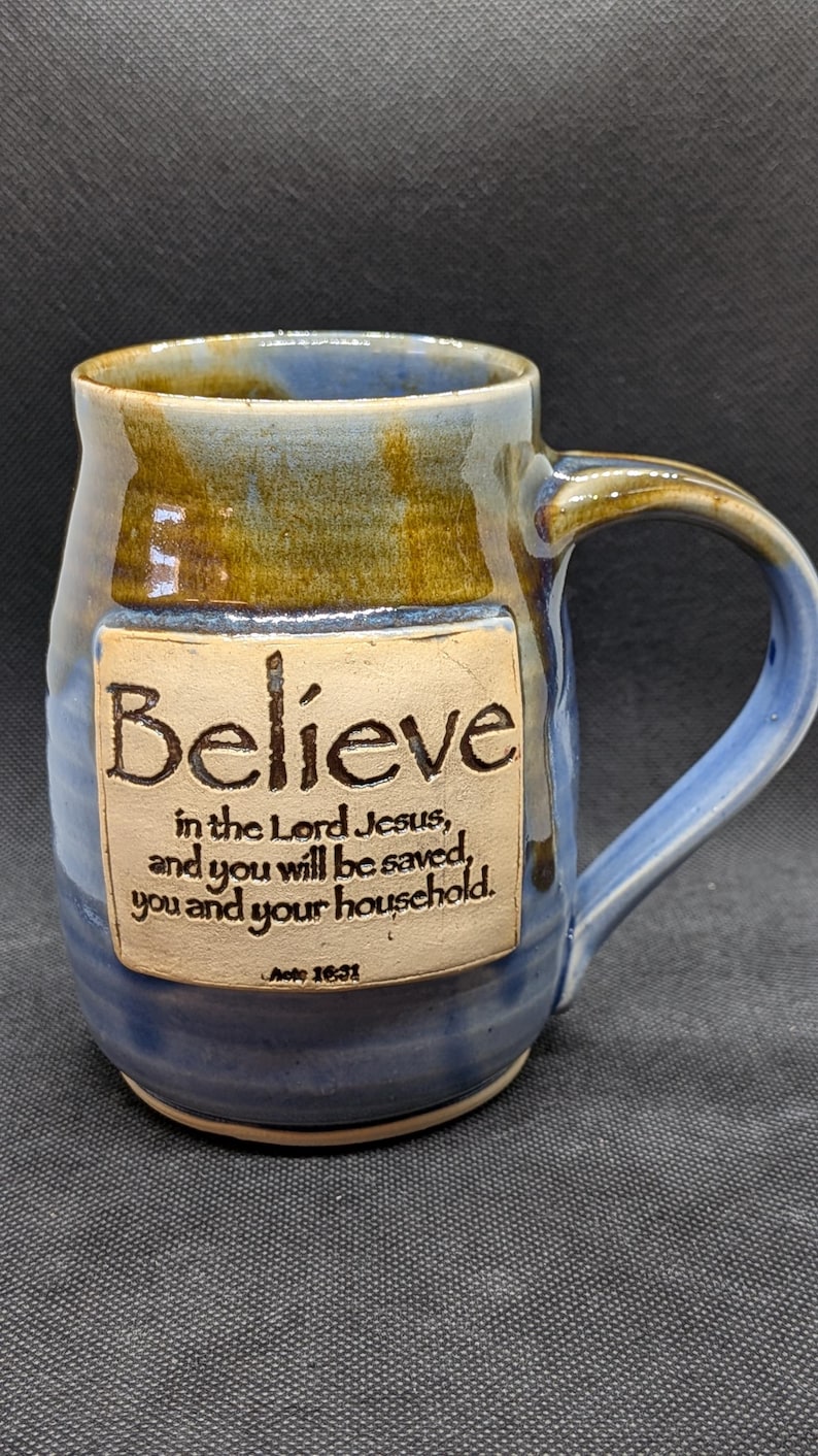 May include: A handcrafted ceramic mug with a blue and brown glaze. The mug features the word "Believe" and a Bible verse. The handle is a curved, sturdy design. A unique piece for coffee or tea.