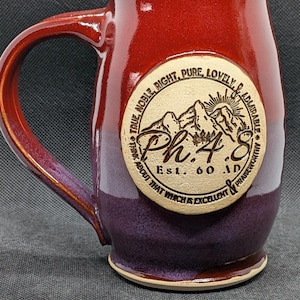 May include: A handcrafted ceramic mug with a burgundy and purple glaze. The mug features a circular emblem with the text "Ph. 4:8" and the words "True, Noble, Right, Pure, Lovely, Admirable, Praise Worthy".