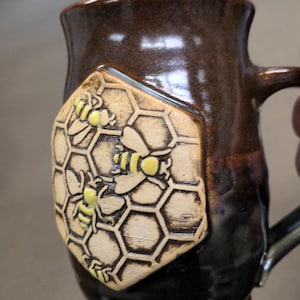 May include: A dark brown ceramic mug featuring a raised, hexagonal-shaped emblem on the front. The emblem displays a textured honeycomb pattern with three small, yellow-painted bees. The mug has a glossy finish and a curved handle on the side.