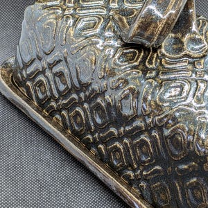 May include: A ceramic butter dish with a lid and a handle. The dish is dark with a textured, geometric pattern in shades of black and gold. The handle is a curved loop.
