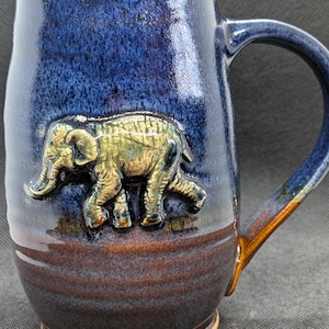 May include: A handcrafted ceramic mug with a blue and brown glaze. An embossed elephant design is featured on the front. The mug has a curved handle and measures approximately 4 inches in height.