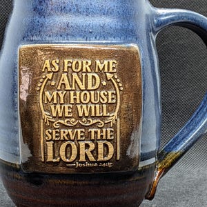 May include: Blue and brown ceramic mug with a handle. The mug features a rectangular, brown plaque with the engraved text "AS FOR ME AND MY HOUSE WE WILL SERVE THE LORD - Joshua 24:15."