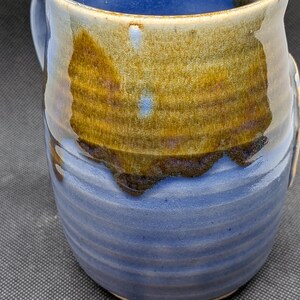 May include: Handmade ceramic mug with a blue interior and exterior, accented by a band of brown and gold glaze. The mug has a rounded shape with a handle and a textured, ridged design on the lower half.