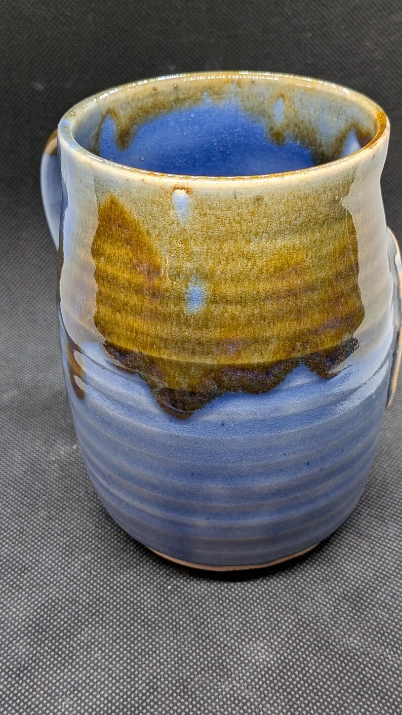 May include: Handmade ceramic mug with a blue interior and exterior, accented by a band of brown and gold glaze. The mug has a rounded shape with a handle and a textured, ridged design on the lower half.