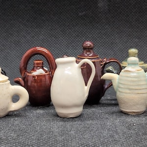 May include: A collection of miniature ceramic teapots and a creamer in various colors, including black, brown, white, and green. The teapots have different shapes and handle designs, showcasing a variety of styles.