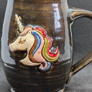 May include: A handmade ceramic mug with a dark brown glaze and a sculpted unicorn design. The unicorn has a cream-colored face, a white horn, and a colorful mane in shades of red, blue, pink, and yellow. The mug has a large handle.