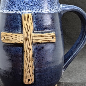 May include: A ceramic mug with a dark blue glaze and a textured rim. A wooden cross is affixed to the front of the mug. The mug has a large handle and a light brown base.