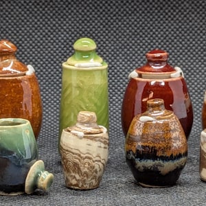 May include: A collection of ceramic jars and containers in various sizes and colors. The jars have lids and are glazed in shades of brown, green, and blue. Some have a marbled effect, while others are solid.