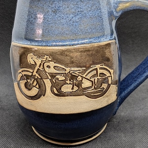 May include: A handcrafted ceramic mug with a blue and brown color scheme. The mug features a detailed relief of a motorcycle. The handle and rim are blue, while the central band showcases the motorcycle design in a contrasting brown.
