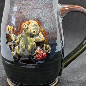 May include: A handmade ceramic mug with a dark brown and black glaze. The mug features a raised relief of a whimsical creature in shades of green and gold, holding a cluster of red berries. The handle is dark green.