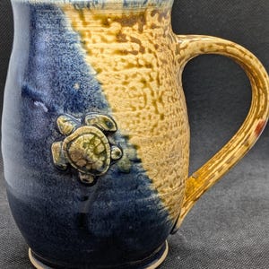 May include: Ceramic mug with a turtle design. The mug features a blue and tan color scheme with a textured finish. A raised turtle is on the side. The handle is tan and the rim is blue.