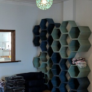 May include: A wall-mounted hexagonal shelf unit with a gradient of blue and green paint. The shelves are empty except for a few blankets on the top shelf. The shelves are in a room with a wooden floor and a window in the background.