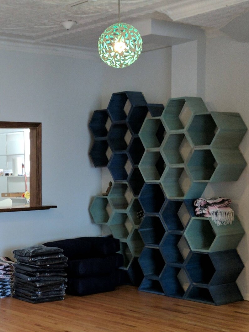 May include: A wall-mounted hexagonal shelf unit with a gradient of blue and green paint. The shelves are empty except for a few blankets on the top shelf. The shelves are in a room with a wooden floor and a window in the background.