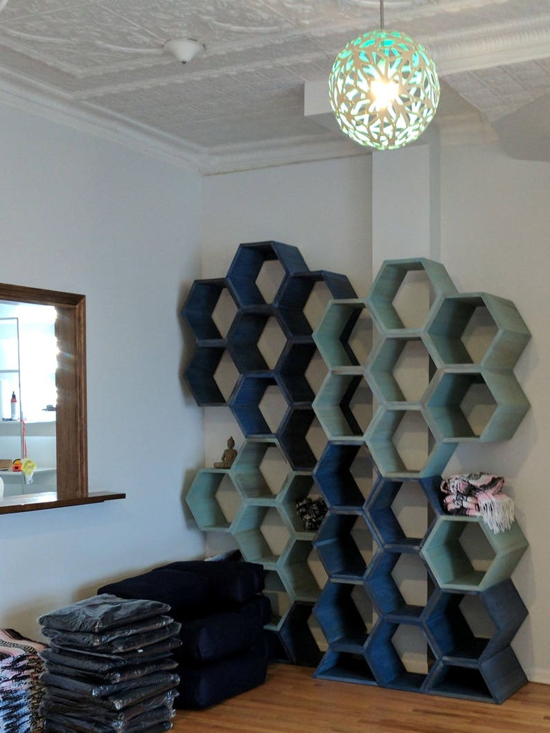 May include: A wall unit with hexagonal shelves in shades of blue and green. The shelves are stacked in a staggered pattern and are empty except for a small Buddha statue on one of the shelves. There are stacks of folded blankets in the foreground.