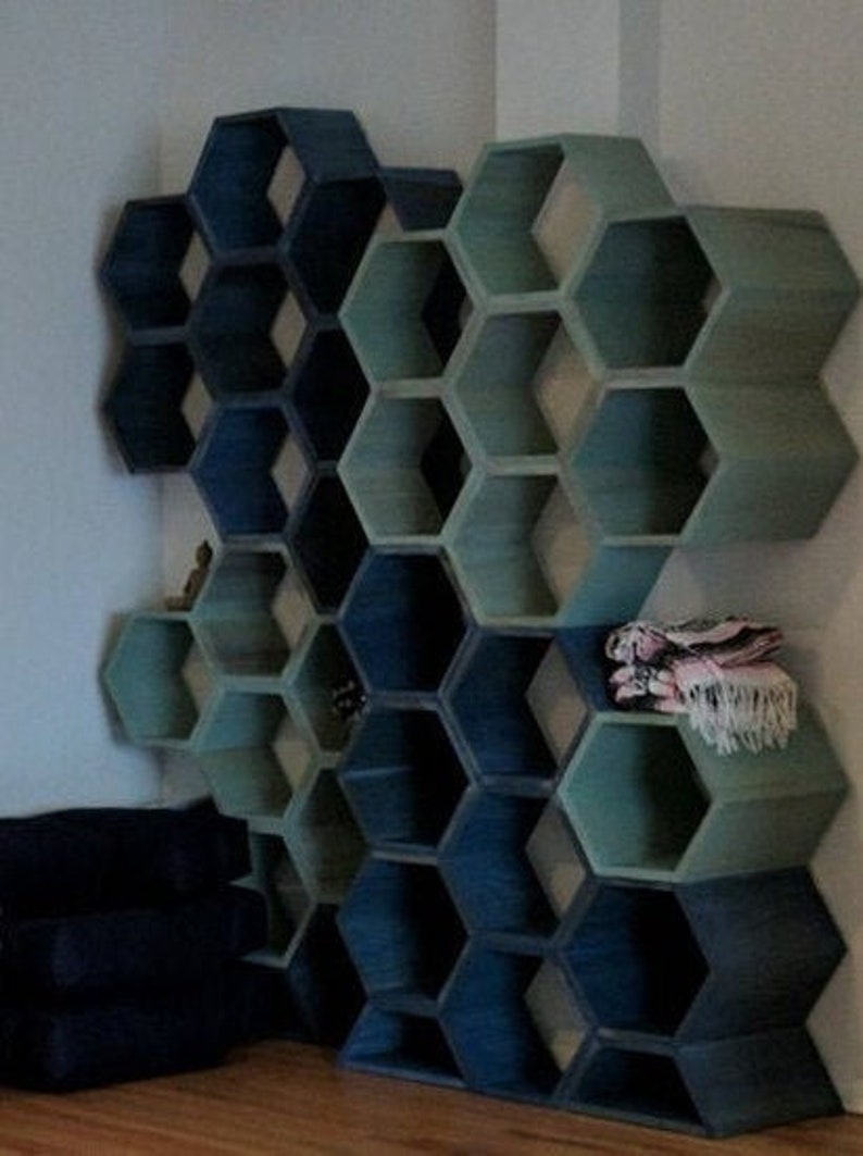 May include: A wall-mounted hexagonal shelf unit made of wood. The shelves are painted in shades of blue and green, creating a gradient effect. The shelves are empty, but there is a small decorative object on one of the lower shelves.