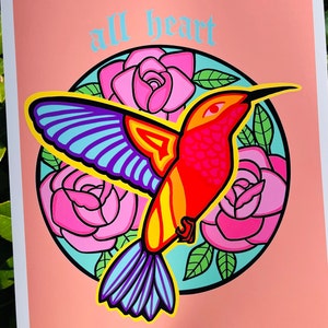 8.5x11 digital illustration featuring a fiery hummingbird in oranges and yellows with a halo of pink roses behind them and the words ‘all heart’ on top of the halo in blue and old English type. The background is a blush orange tone.