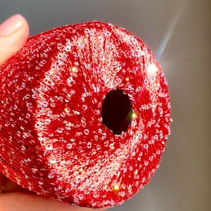 May include: A close-up of a red, sequined yarn ball with a hole in the center. The yarn is a vibrant red color, and the sequins add a sparkling effect. The yarn ball is held in a hand.