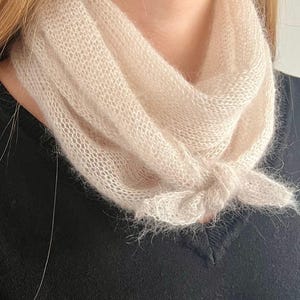 Könnte beinhalten: Ein weicher, weißer, gestrickter Schal mit einem zarten, offenen Muster. Der Schal wird um den Hals getragen und in einem lockeren Knoten gebunden.