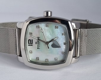 May include: A silver Skagen watch with a white mother-of-pearl face and diamond accents. The watch has a square face and a mesh strap.