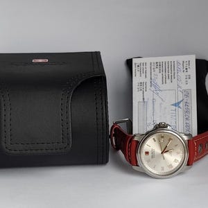 May include: A silver-toned wristwatch with a red leather band and a silver face, next to a black leather watch case and a white document. The watch has Roman numeral hour markers and a date display.