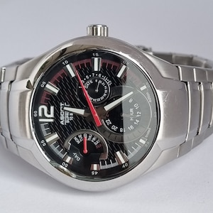 May include: A silver-toned men's wristwatch with a black face and red accents. The watch features a date display, day of the week display, and a 24-hour display. The watch has a stainless steel strap.