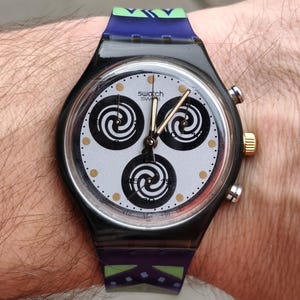May include: A black Swatch watch with a silver face featuring three black spiral designs. The watch has gold and silver accents, and a patterned blue and green strap. The watch is on a wrist.