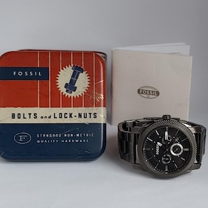 May include: A black Fossil watch with a metal strap, alongside a red, white, and blue tin that says "BOLTS and LOCK-NUTS". A booklet with the Fossil logo is also visible.