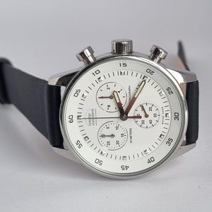 May include: A silver chronograph wristwatch with a white face and black leather strap. The watch has a date window, a chronograph function, and a 50-metre water resistance rating. The watch face features the text "Vivadent" and "Chronograph".