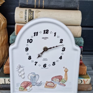 Pode incluir: Relógio de cerâmica branca com mostrador preto e ponteiros pretos. O relógio tem um design decorativo de comida e um moinho de pimenta. O mostrador do relógio diz "seratec time" e "QUARTZ  klockmall  MADE IN GERMANY".
