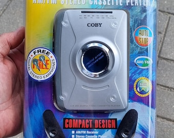 May include: A silver and black AM/FM stereo cassette player with a compact design. The player has a digital tuner with a display showing the frequency. The packaging includes a pair of free digital stereo earphones.