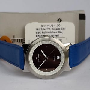 May include: A silver-toned Junghans Solar TEC watch with a dark brown face and blue leather band. The watch has white hands and markers, and the date is displayed at the 6 o'clock position. The watch is displayed with a product information tag.